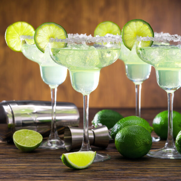 Four glasses of freshly made Margarita cocktail decorated with limes on wooden table with shaker and limes.